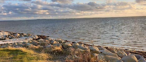 A coastal landscape with a rocky shoreline, grassy areas, and a calm sea under a partly cloudy sky.