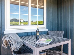 A wooden table with a bottle and a magazine on it, outside a building with a window.