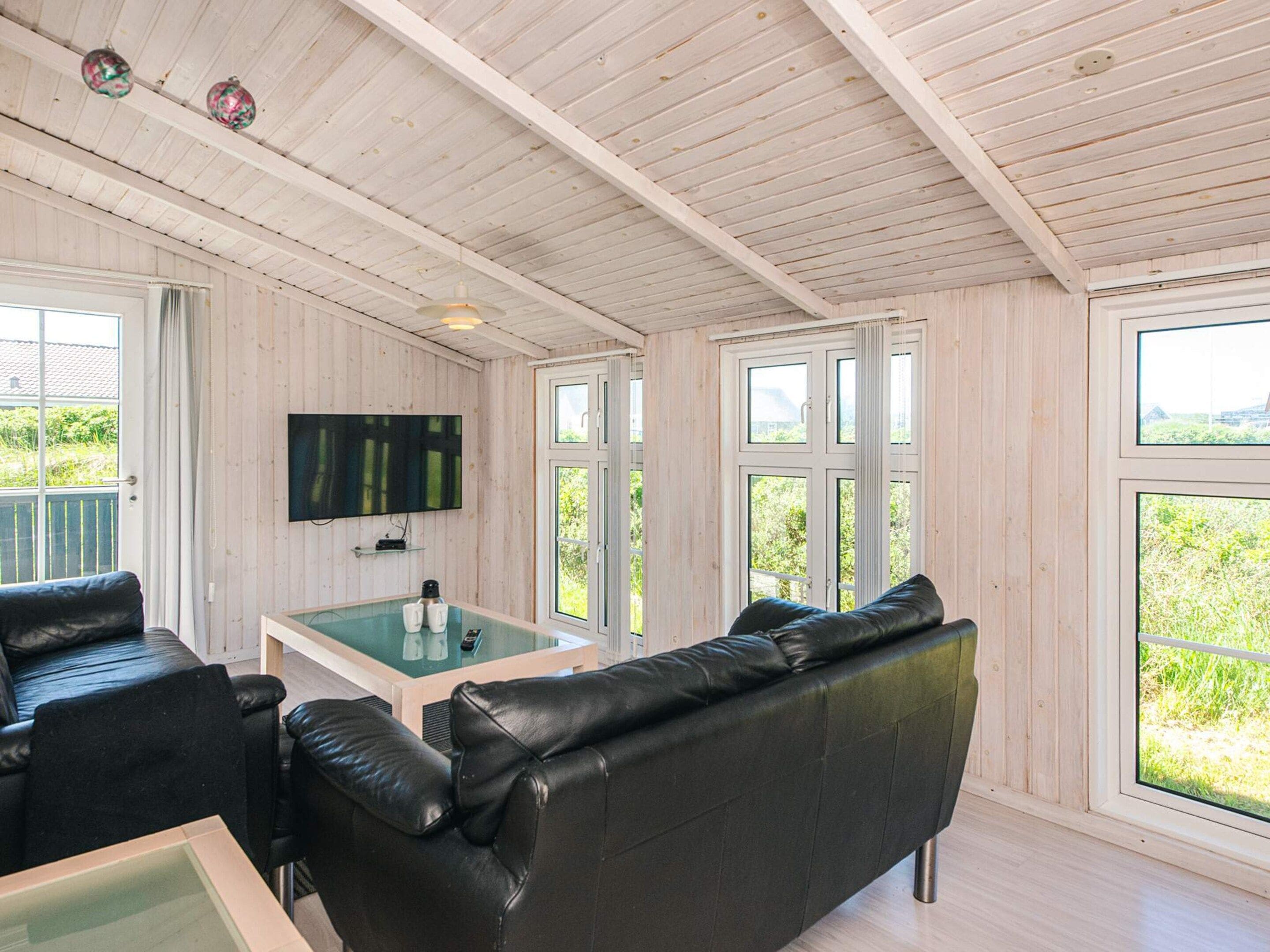 Une pièce dotée d’un plafond en bois, d’un canapé en cuir noir, d’une table basse en verre, d’un téléviseur à écran plat fixé au mur et de grandes fenêtres donnant sur un paysage verdoyant.