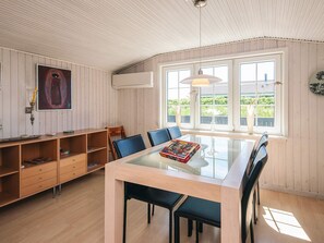 Een kamer met een eettafel met glazen blad, blauwe stoelen, een houten boekenkast en een groot raam met uitzicht op groen.