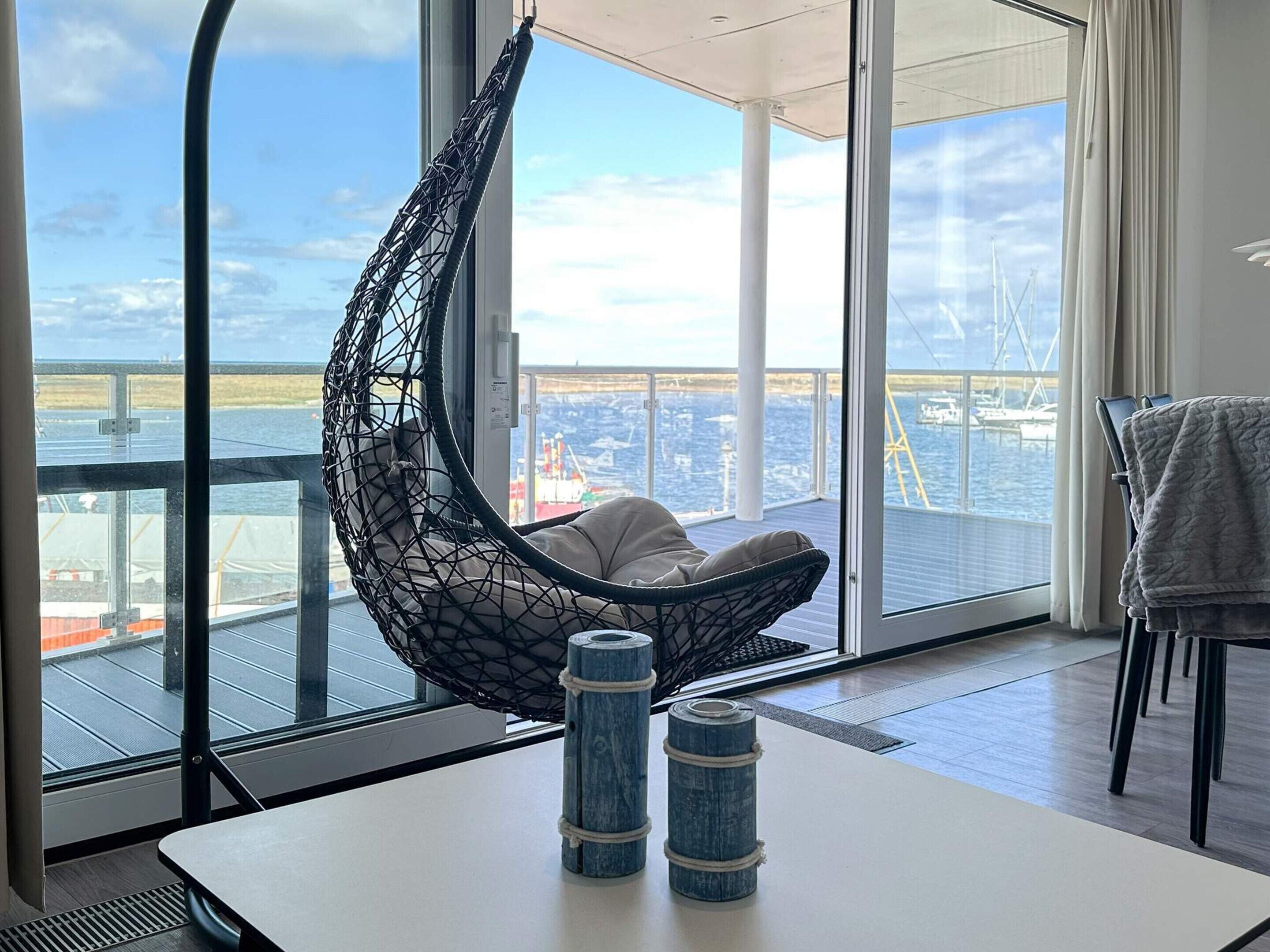 Ein Zimmer mit einem Hängekorbstuhl, einem Tisch mit zwei blauen Behältern und Blick auf Meer und Schiffe durch große Fenster.