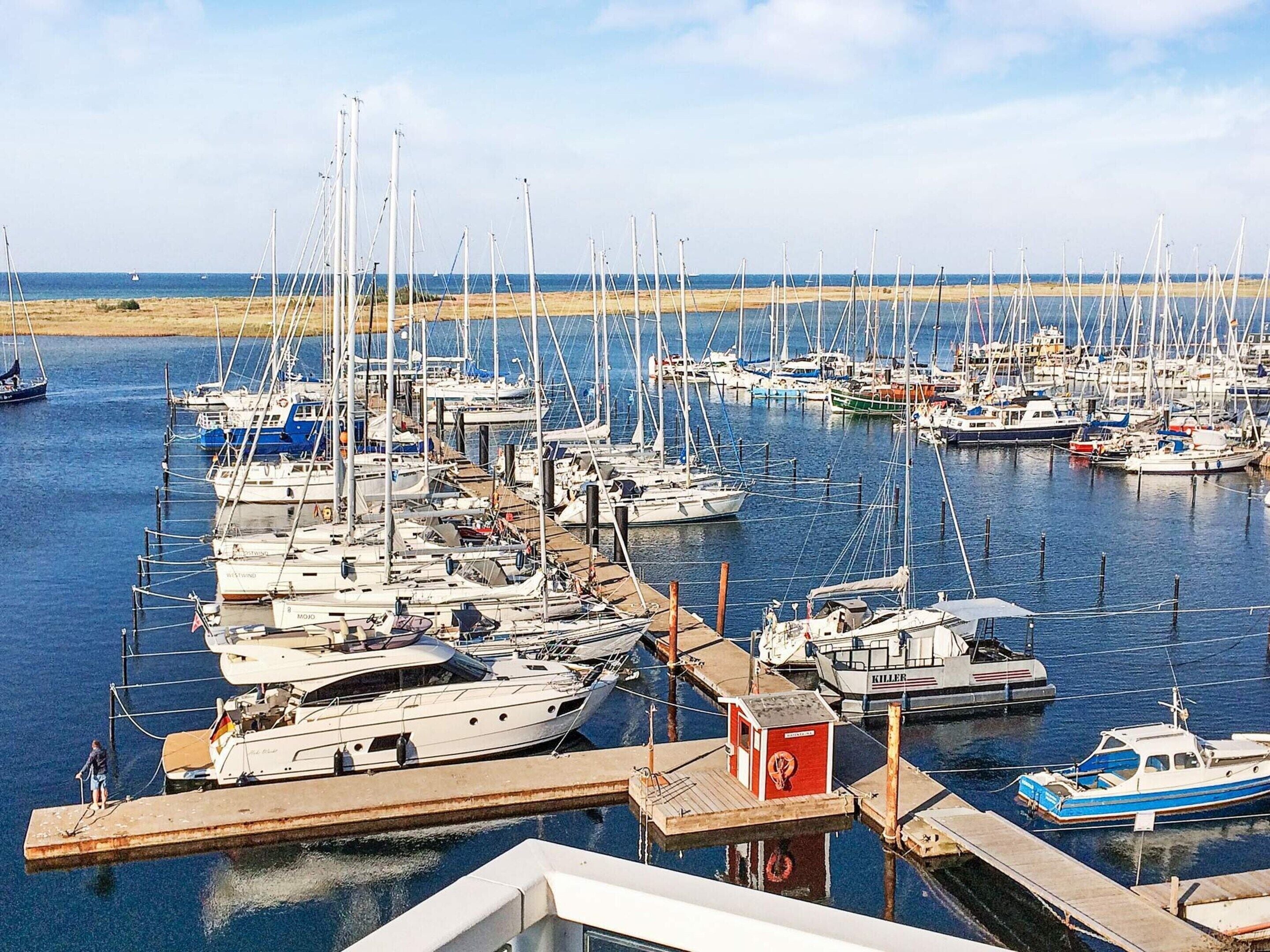 A marina with numerous boats docked, a small red building, and a sandy beach in the background.