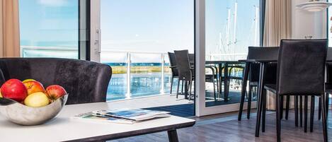 A room with a dining area, a table with a bowl of fruit, and a view of the sea.