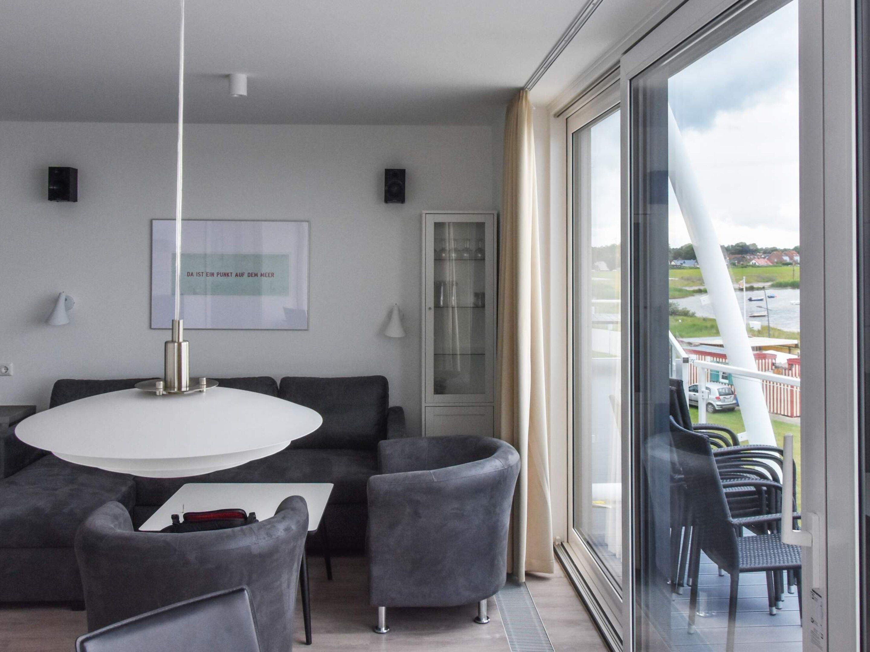 Un moderno salón con una mesa redonda, sofás grises, un aparador de vidrio y vistas a un cuerpo de agua y edificios.