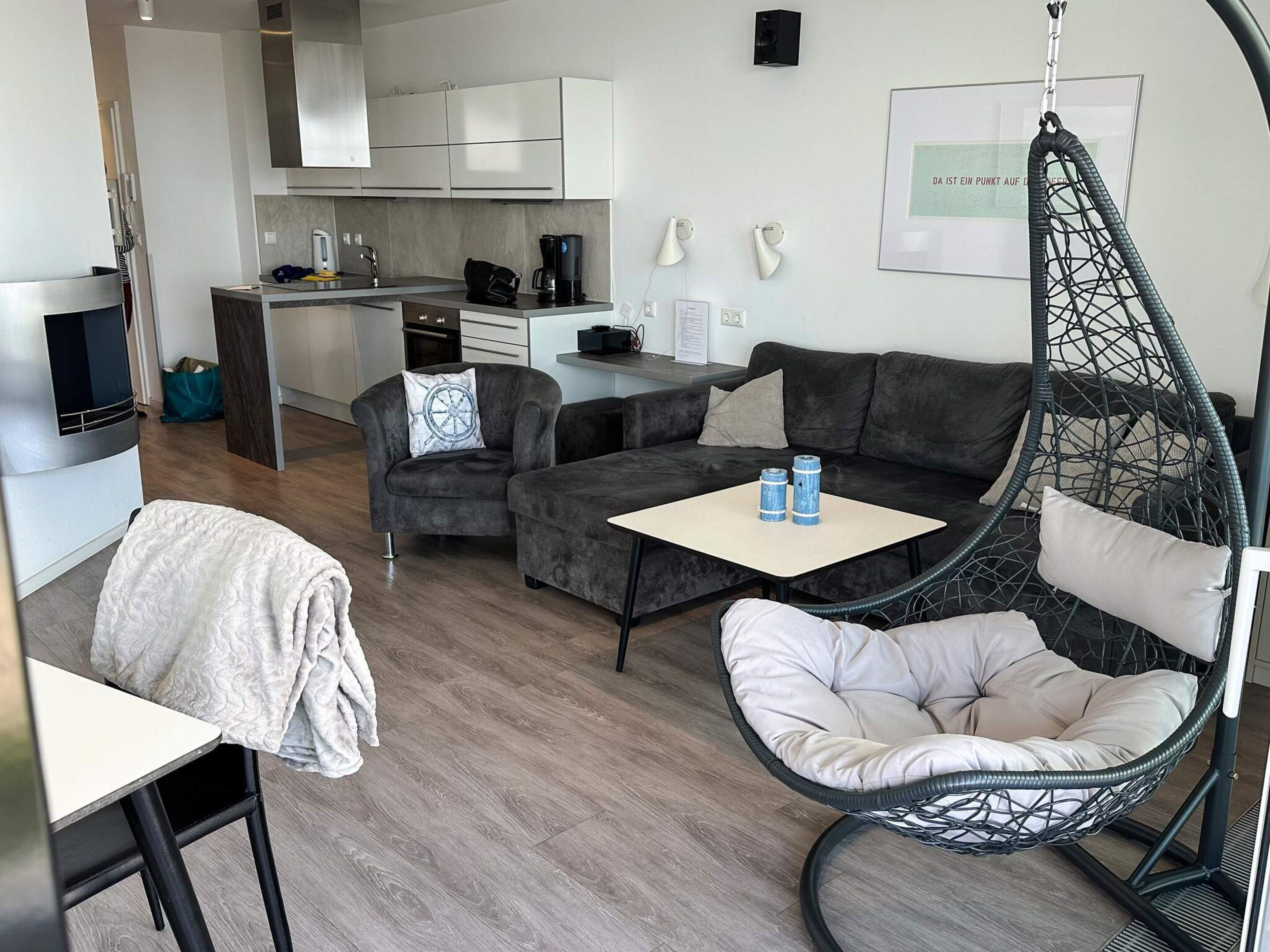 A modern living room with a sofa, a hanging chair, and a small table. There is a kitchen in the background with white cabinets and a coffee machine.