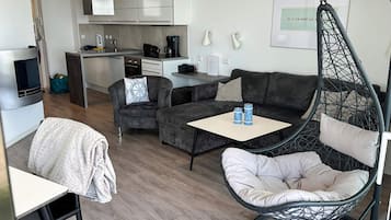 A modern living room with a sofa, a hanging chair, and a small table. There is a kitchen in the background with white cabinets and a coffee machine.