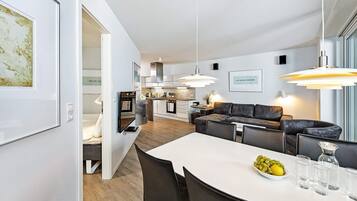 Eine moderne Küche mit weißen Schränken, ein Essbereich mit einem weißen Tisch und schwarzen Stühlen sowie ein Wohnbereich mit einem dunklen Sofa.