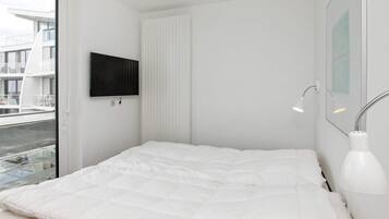 Ein modernes Schlafzimmer mit einem großen Bett, einem an der Wand befestigten Fernseher, einem Fenster mit Blick auf Gebäude und einer Nachttischlampe.