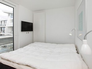 A modern bedroom with a large bed, a mounted TV, a window with a view of buildings, and a bedside lamp. - Holiday Home in Wendtorf (Schönberg)