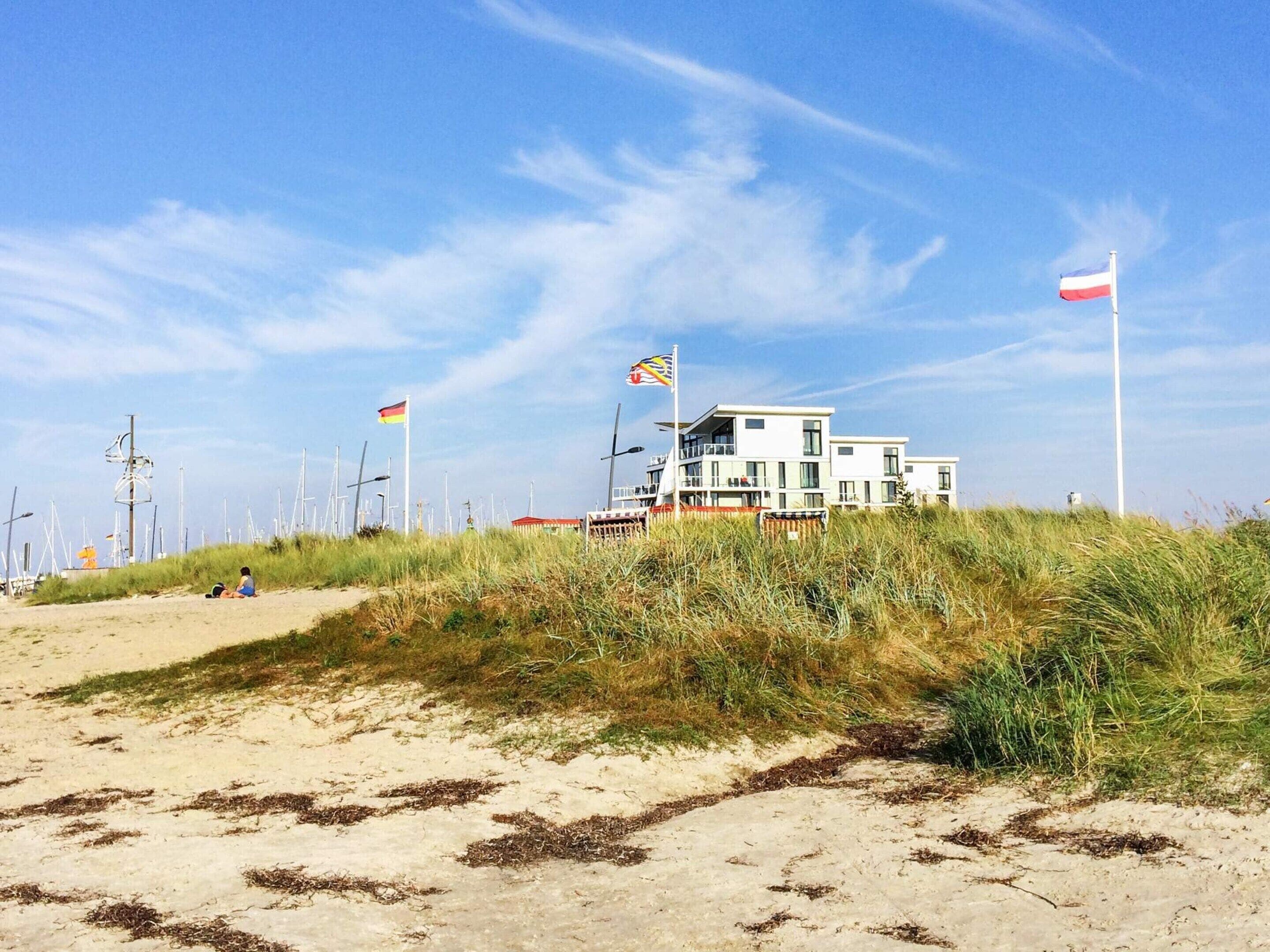 A coastal building with multiple flags, surrounded by dunes and wind turbines.