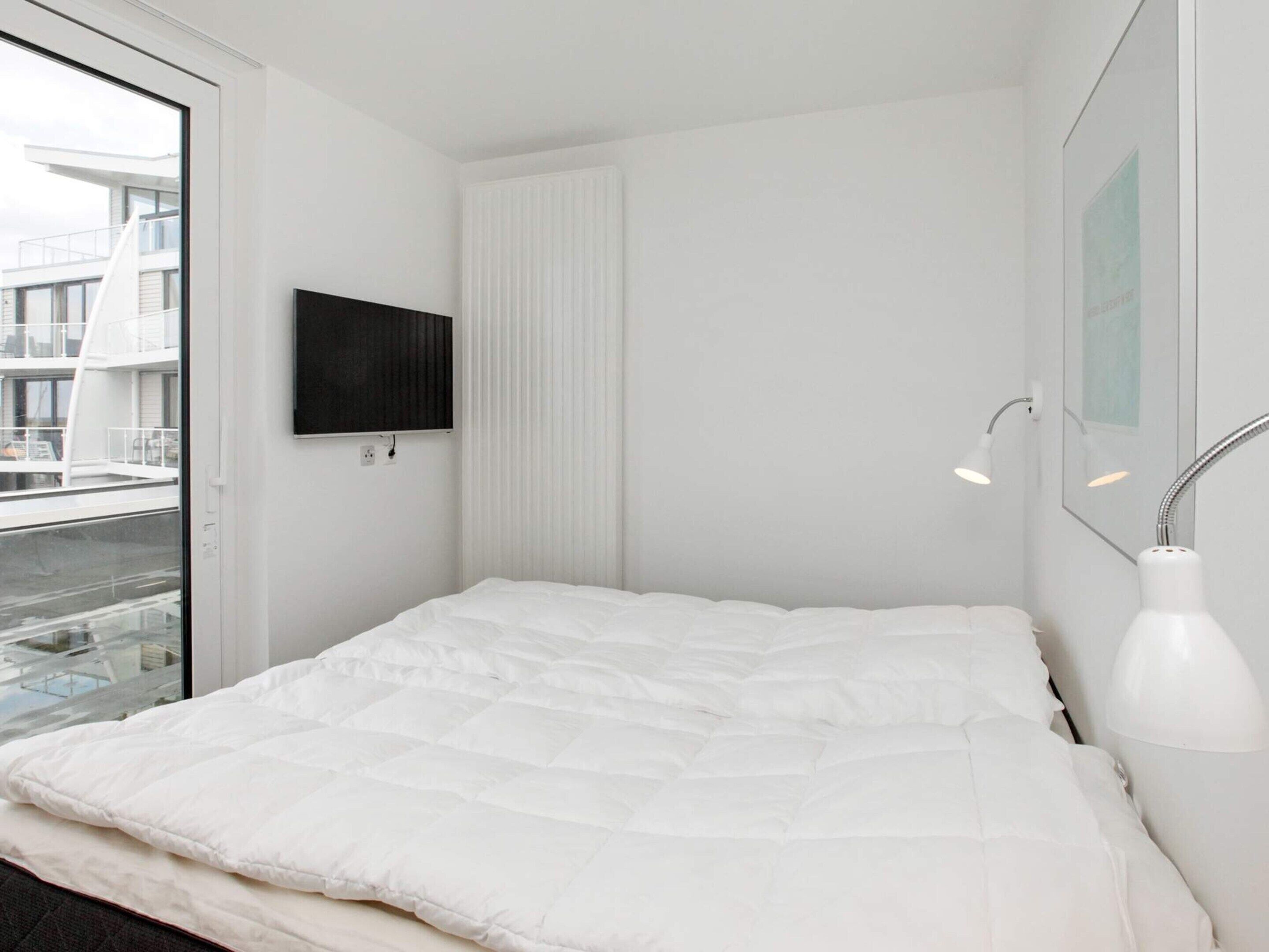 A modern bedroom with a large bed, a mounted TV, a bedside lamp, and a view of the outside.
