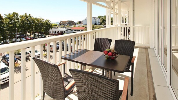 A balcony with a table and chairs, a bowl of fruit, and a view of the street and buildings. - Holiday Home in Grossenbrode (Grossenbrode)