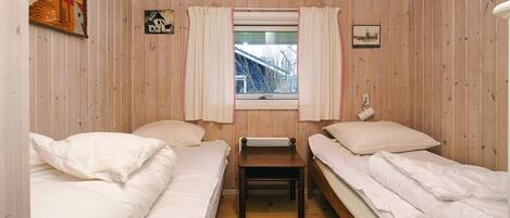 A room with two single beds, a wooden bench, a window with curtains, and a view of a building outside.