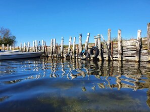 Ruhige Wasserfläche mit Holzpfählen und Reifen, ein Boot ist am linken Ufer vertäut, und ein klarer, blauer Himmel darüber.