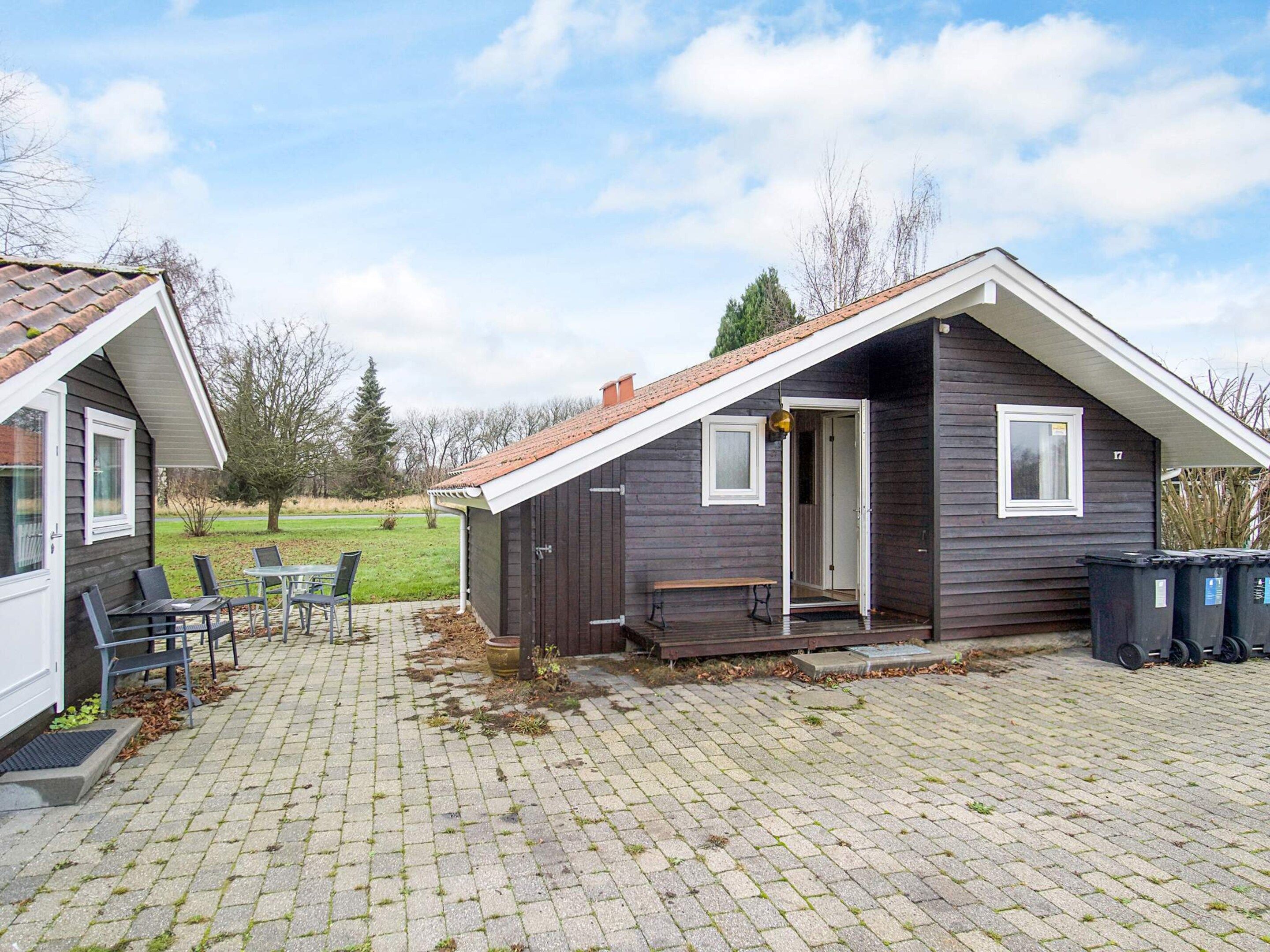 Une petite cabane en bois sombre, avec un toit en pente, une terrasse avec une table et des chaises, et une cour verdoyante en arrière-plan.