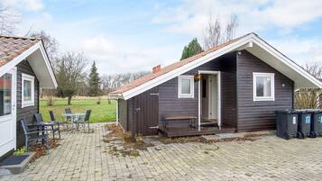 Une petite cabane en bois sombre, avec un toit en pente, une terrasse avec une table et des chaises, et une cour verdoyante en arrière-plan.