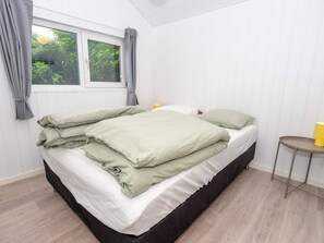 A single bed with a white mattress and green bedding, a small bedside table with a yellow lamp, and a window with curtains. - 4 Star Holiday Home in Harboore (Harboore)