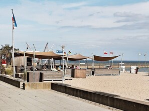 Un muelle frente a la playa con piso de madera, zonas de descanso a la sombra y vistas al océano.
