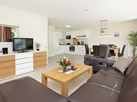 A modern living room with a sofa, a coffee table with a vase of flowers, a television, and a kitchen in the background.