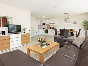 A modern living room with a sofa, a coffee table with a vase of flowers, a television, and a kitchen in the background. - Holiday Home in Großenbrode (Grossenbrode)