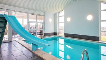 En indendørs swimmingpool med blå rutsjebane, klare glasvægge og udsigt til det fri.