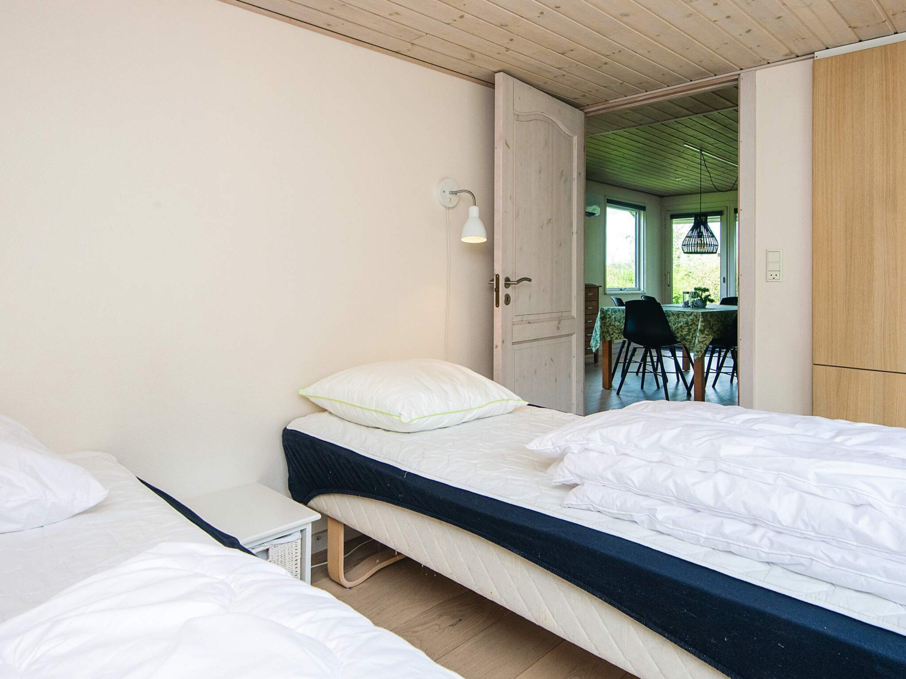 Une chambre avec deux lits simples, une petite table de chevet, un luminaire et une porte ouverte donnant sur un coin repas avec une table et des chaises.