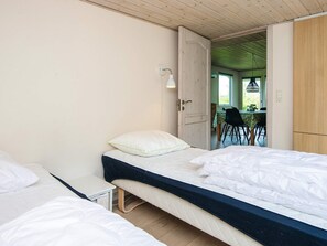 Un dormitorio con dos camas individuales, una mesita de noche pequeña, una lámpara y una puerta abierta que da a un comedor con una mesa y sillas.