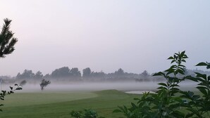 Un campo da golf avvolto nella nebbia, con una bandierina, alberi e un cielo sereno.