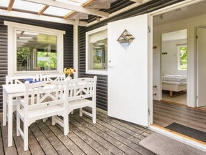 Een houten terras met een witte tafel en stoelen, een deur naar een slaapkamer met een bed, en een klein raam met uitzicht op groen.