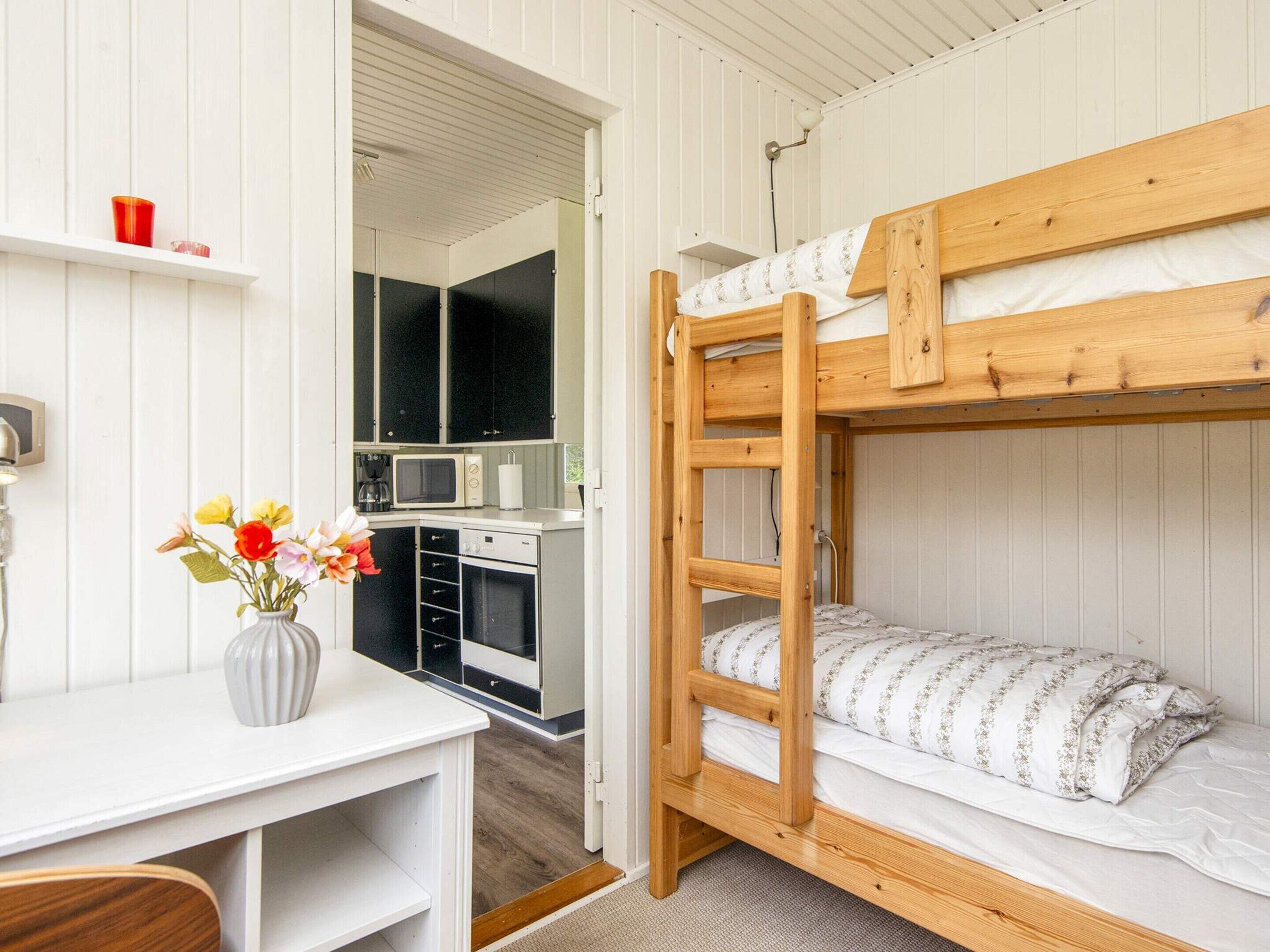 A bunk bed with a wooden frame and white bedding, a small kitchenette with a microwave and a sink, and a desk with a vase of flowers.