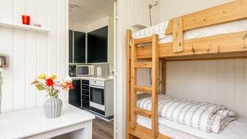 A bunk bed with a wooden frame and white bedding, a small kitchenette with a microwave and a sink, and a desk with a vase of flowers.