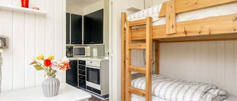 A bunk bed with a wooden frame and white bedding, a small kitchenette with a microwave and a sink, and a desk with a vase of flowers.