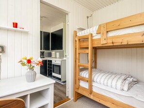 Een stapelbed met houten frame en witte beddengoed, een kleine kitchenette met een magnetron en spoelbak, en een bureau met een bloevas.