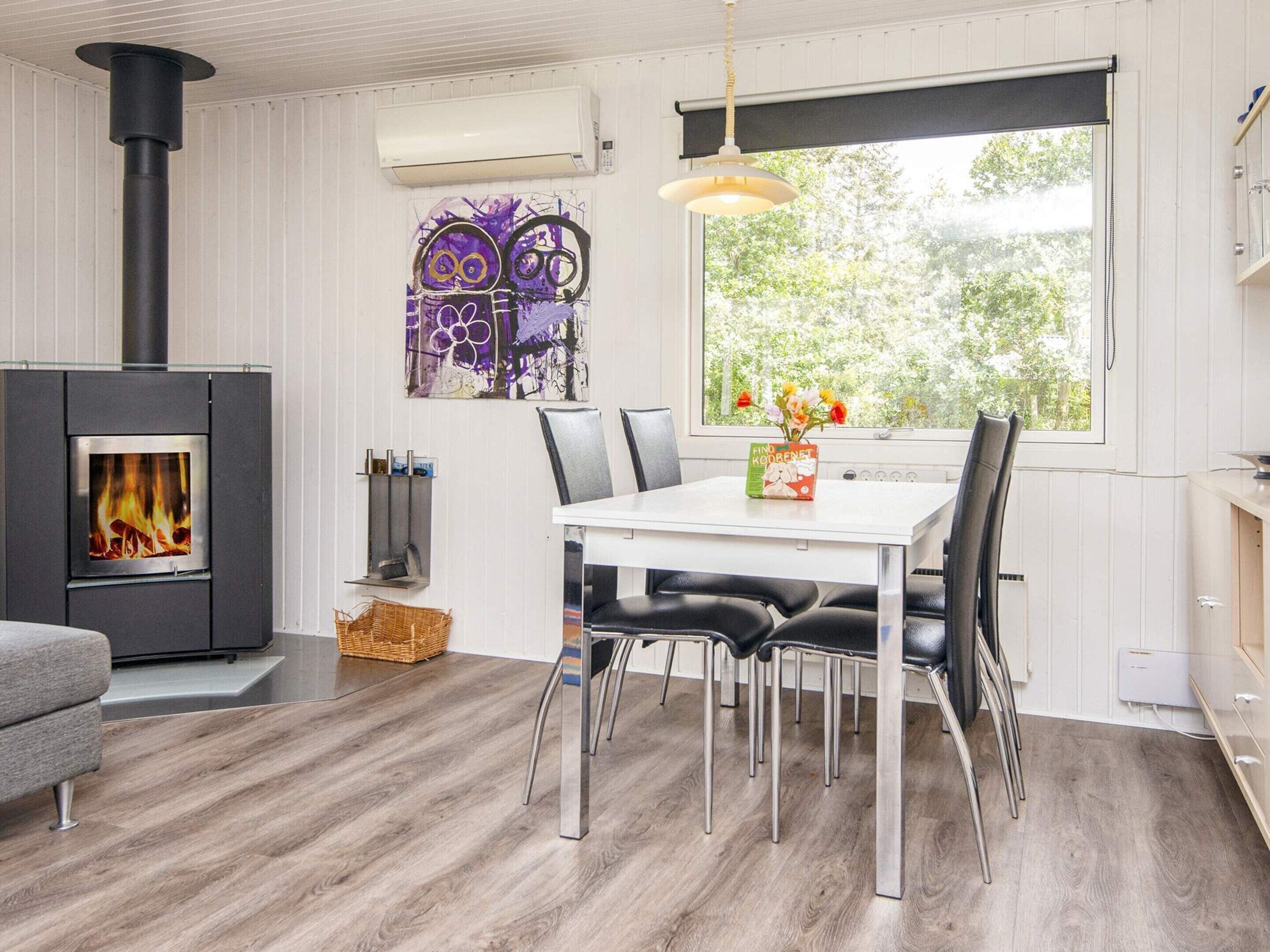 A modern interior with a wood-burning stove, a dining table with chairs, and a large window with a view of greenery.
