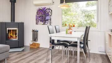 A modern interior with a wood-burning stove, a dining table with chairs, and a large window with a view of greenery.
