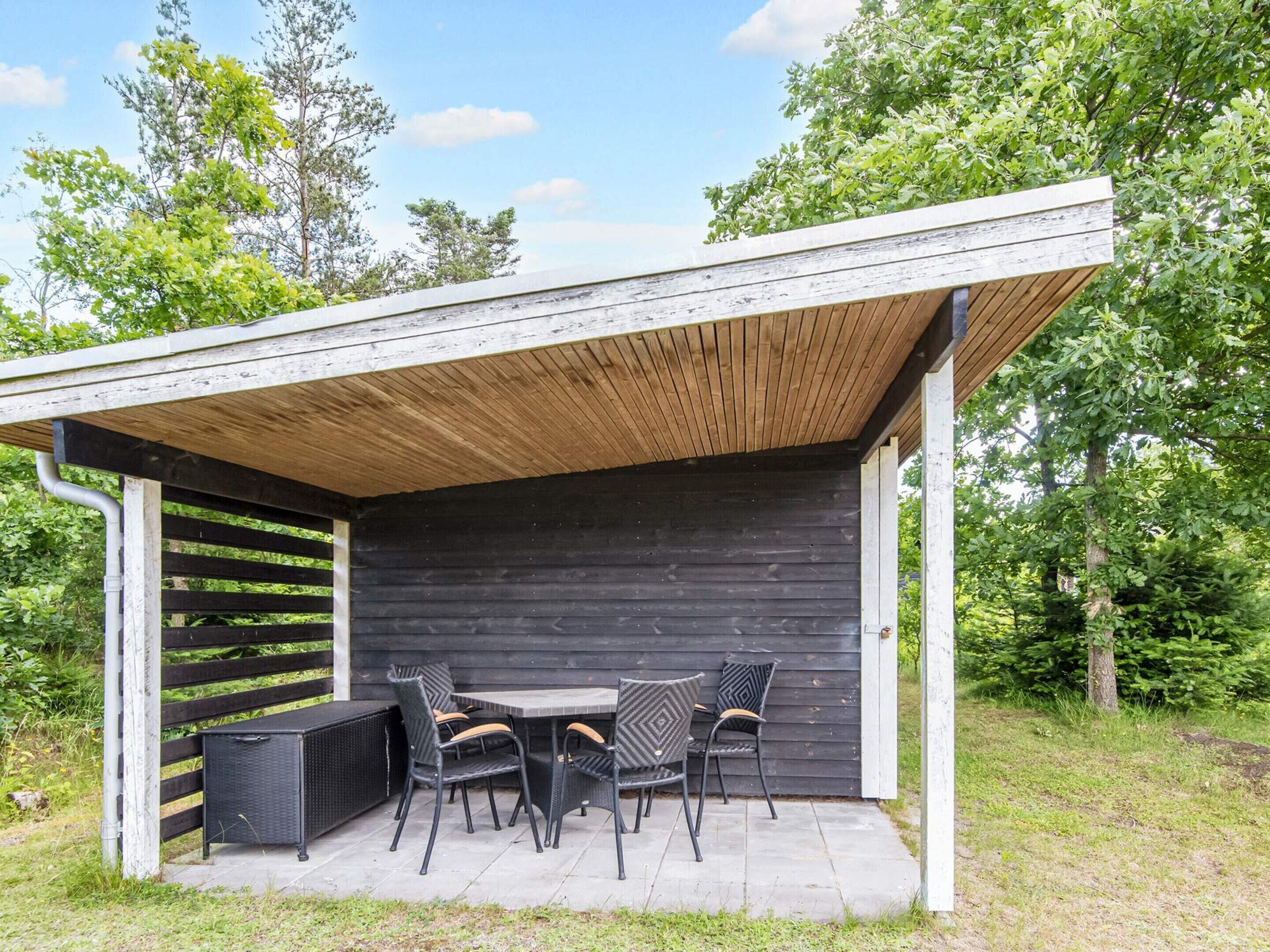 A covered outdoor seating area with a table and chairs, surrounded by greenery.