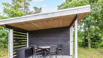 A covered outdoor seating area with a table and chairs, surrounded by greenery.