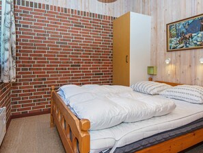 A bedroom with a brick-patterned wall, a wooden bed frame, a green wall-mounted lamp, and a painting of a forest scene.