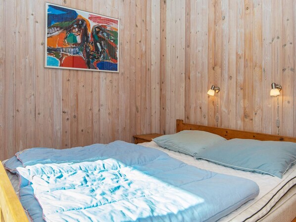 A bedroom with a wooden wall, a bed with blue bedding, a colorful painting, and two wall-mounted lamps.