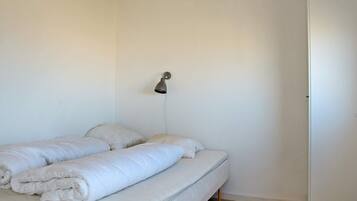 Una cama individual con ropa de cama blanca en una habitación con paredes blancas y suelo de madera.