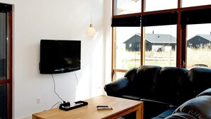 A modern living room with a flat-screen TV mounted on the wall, a leather sofa, a wooden coffee table, and a view of houses through large windows.