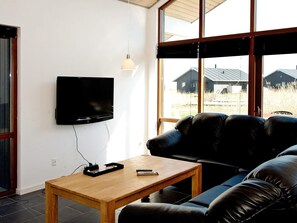 A modern living room with a flat-screen TV mounted on the wall, a leather sofa, a wooden coffee table, and a view of houses through large windows. - 8 Person Holiday Home in Ulfborg (Ulfborg)