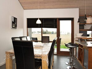 Uma sala de jantar com mesa e cadeiras, uma cozinha com bancada e eletrodomésticos, e vista para o exterior através de uma porta deslizante.