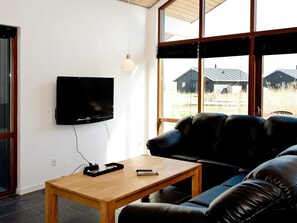 A living room with a flat-screen TV mounted on the wall, a wooden coffee table, a black leather sofa, and a large window with a view of houses.