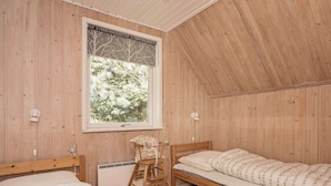 A room with wooden walls and a sloped ceiling, featuring two beds, a chair, and a window with a view of trees.