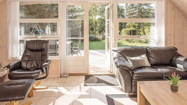 A cozy living room with a leather sofa, a chair, and a coffee table. There are large windows with a view of a garden.