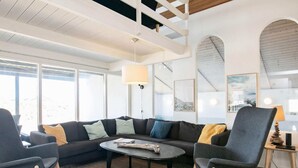 A modern living room with a sectional sofa, two armchairs, a coffee table, and a lamp. Large windows provide natural light and a view of the outdoors.