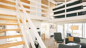 A spacious loft-style room with a wooden staircase, a cozy seating area with a sofa and armchairs, and a large window providing natural light.