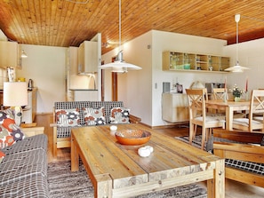 A cozy living room with a wooden table, sofa, and chairs, a kitchen area with cabinets and a dining table, and a ceiling with wooden beams. - Rustic Holiday Home in Asnæs near Sea (Asnaes)