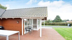 Een overdekt buitengebied met een houten terras, een barbecue en een zithoek met stoelen en een tafel.